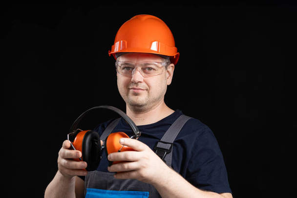Builder σε ένα προστατευτικό κράνος με ακουστικά ακύρωσης θορύβου στα χέρια του σε μαύρο φόντο, αντίγραφο χώρο για κείμενο. - Φωτογραφία, εικόνα