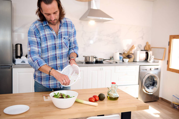 Auténtico retrato de un hombre caucásico guapo, cocinero poniendo verduras secas y hierbas en un tazón blanco mientras cocina ensalada saludable en la cocina casera. Concepto de alimentación saludable y dieta. - Foto, imagen