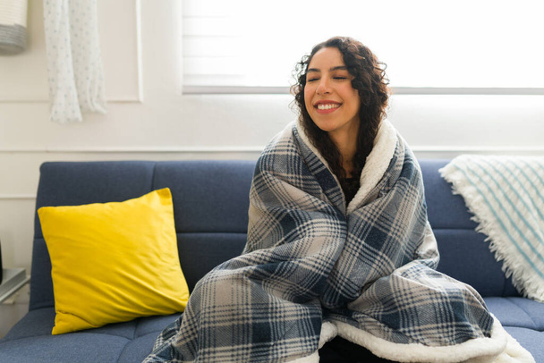 Mujer latina feliz con el pelo rizado sonriendo calentándose con una hermosa manta y relajándose en el sofá disfrutando del clima frío  - Foto, imagen