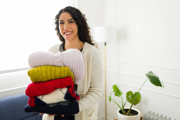 Mujer joven mexicana con el pelo rizado sonriendo mientras saca la ropa de invierno durante el clima frío mirando feliz en la sala de estar - Foto, Imagen