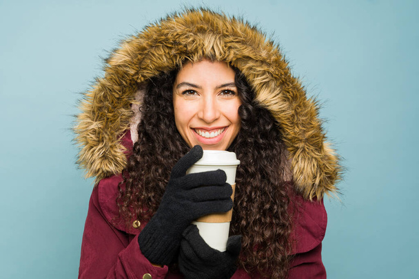 Büyük bir kış ceketi giymiş İspanyol güzeli genç bir kadın gülümseyerek mutlu görünüyor soğuk havada kahve içip eğleniyor. - Fotoğraf, Görsel