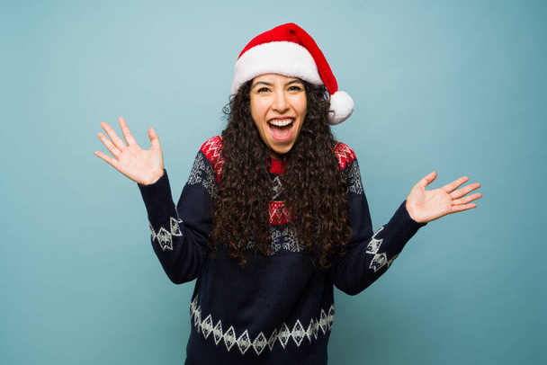 Femme latine excitée criant regardant surpris et heureux au sujet des vacances portant un chapeau de Père Noël et pull de Noël - Photo, image