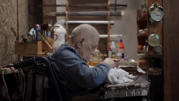 Senior gray haired male master in a wheelchair is working at his workshop sharpening japanese swords blade - Felvétel, videó