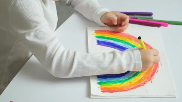 Un niño pequeño dibuja un arco iris con rotuladores en un cuaderno de bocetos mientras está sentado en una mesa. - Metraje, vídeo