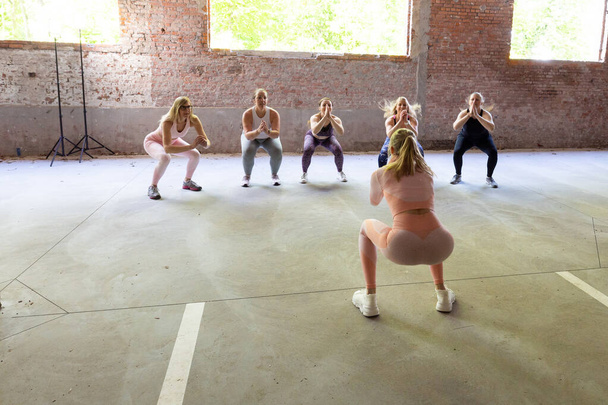 Um grupo diversificado de mulheres, cada uma com seu tipo de corpo único, entusiasticamente se envolver em um treino dinâmico liderado por um personal trainer dentro de casa em um ginásio de fitness, realizando saltos e burpees. Mulheres Diversas - Foto, Imagem