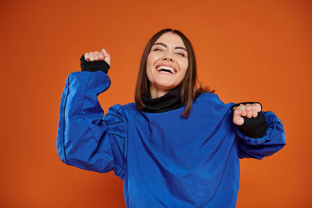ενθουσιασμένοι νεαρή γυναίκα με τρυπημένη μύτη gesturing και χαμογελώντας σε πορτοκαλί φόντο, μπλε φούτερ - Φωτογραφία, εικόνα