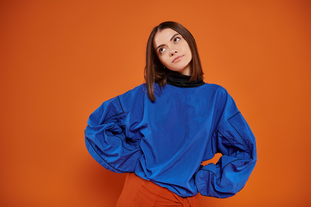 displeased young woman with pierced nose standing with hands on hips on orange backdrop, look away - Photo, Image