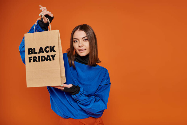 brunette young woman in autumn attire holding shopping bag with black friday letters on orange - Photo, Image