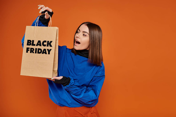 amazed young woman in autumn attire looking at shopping bag with black friday letters on orange - Photo, Image