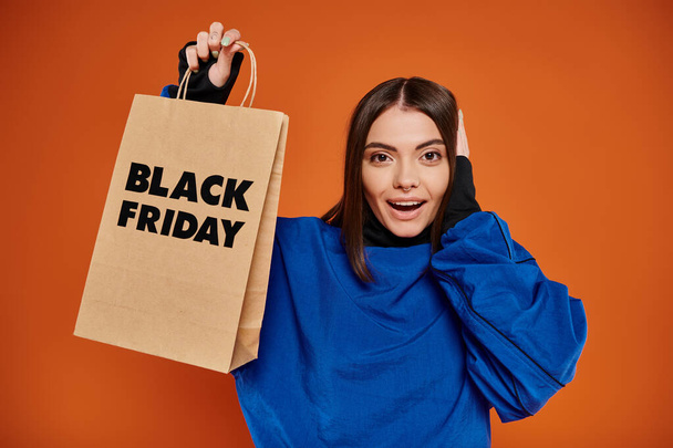 aufgeregte junge Frau in Herbstkleidung mit Einkaufstasche mit schwarzen Freitagsbuchstaben auf Orange - Foto, Bild