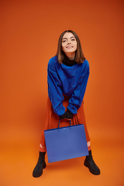 オレンジ色のバックドロップ,ブラックフライデーのコンセプトにショッピングバッグと立っている秋の服装の陽気な女性 - 写真・画像