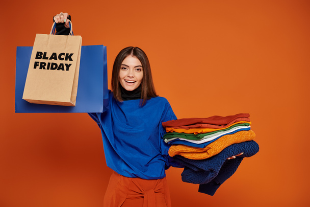 joyful woman holding shopping bags and stack of clothes on orange backdrop, black friday concept - Photo, Image