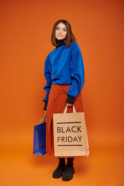 fiatal vidám nő őszi ruha gazdaság bevásárló táskák narancssárga háttér, fekete pénteki értékesítés - Fotó, kép