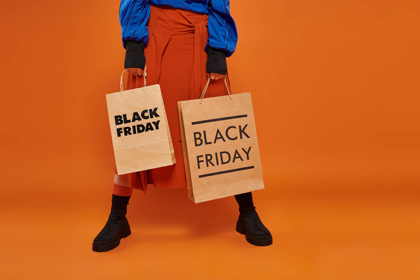 オレンジ色の背景にショッピングバッグを保持する秋のスカートとブーツでクロップされた女性, 黒の金曜日 - 写真・画像