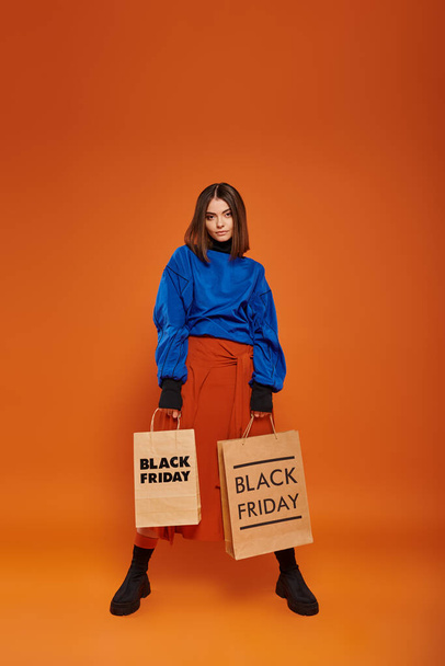 pełna długość, brunetka kobieta w jesiennym stroju trzymając torby na zakupy na pomarańczowym tle, czarny piątek - Zdjęcie, obraz