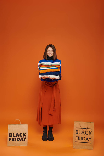 freudige Frau hält Stapel warmer Kleidung in der Nähe von Einkaufstüten mit schwarzen Freitagsbuchstaben auf orange - Foto, Bild