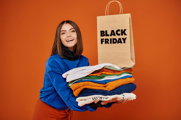 веселая женщина держит стопку теплой одежды и сумку для покупок на оранжевом фоне, черная пятница - Фото, изображение