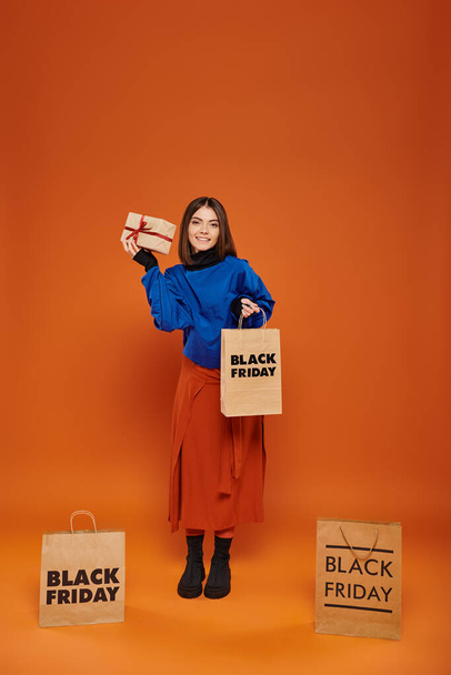 χαρούμενη γυναίκα κρατώντας τυλιγμένο δώρο και τσάντα ψώνια σε πορτοκαλί φόντο, μαύρο πωλήσεις Παρασκευή - Φωτογραφία, εικόνα