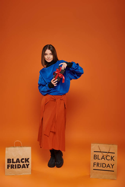 happy woman holding wrapped present near shopping bags on orange backdrop, black friday sales - Photo, Image