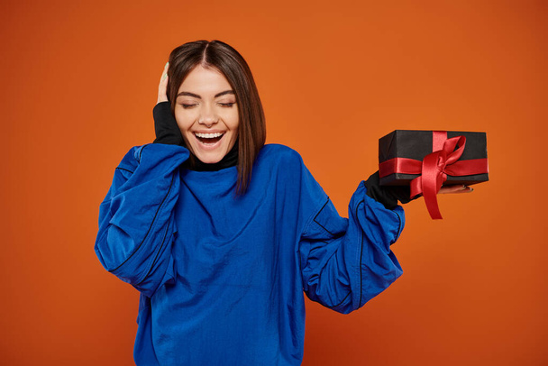 happy woman with brunette hair holding wrapped present on orange background, black friday - Photo, Image