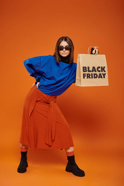 mujer con estilo en gafas de sol de moda con bolsa de compras con letras de viernes negro sobre fondo naranja - Foto, imagen