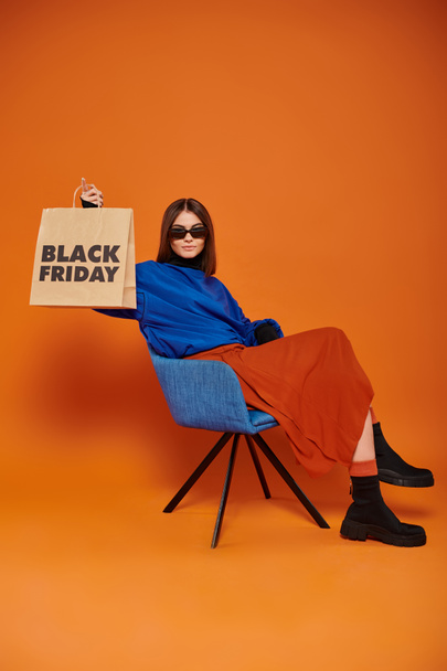 woman in sunglasses and autumnal attire holding black friday shopping bag and sitting on armchair - Photo, Image