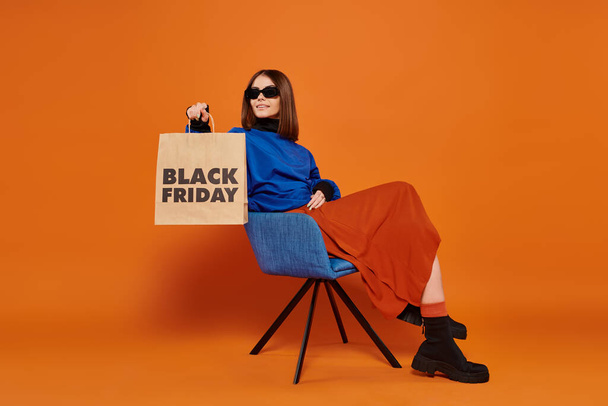 szczęśliwa kobieta w stylowych okularach przeciwsłonecznych gospodarstwa czarny piątek torba na zakupy i siedzi na fotelu - Zdjęcie, obraz