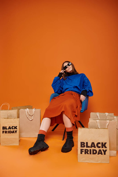 κουρασμένη γυναίκα με κομψά γυαλιά ηλίου κάθεται στην πολυθρόνα κοντά σε μαύρες σακούλες για ψώνια Παρασκευή σε πορτοκαλί - Φωτογραφία, εικόνα