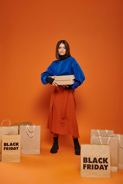 happy woman holding carton box and standing near shopping bags with black friday letters, orange - Photo, Image