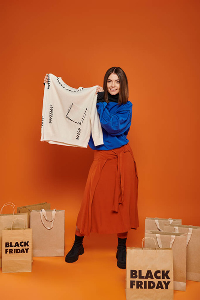 ευτυχισμένη γυναίκα με κοντά μαλλιά κρατώντας λευκή μπλούζα κοντά σε τσάντες για ψώνια με μαύρα γράμματα της Παρασκευής - Φωτογραφία, εικόνα