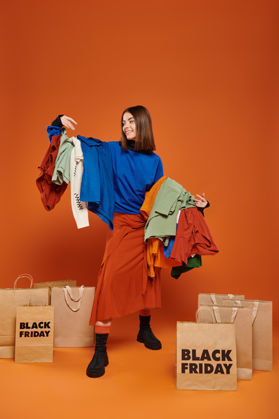 mulher feliz com cabelo morena segurando roupas outonais coloridas, perto de sacos de compras sexta-feira preto - Foto, Imagem
