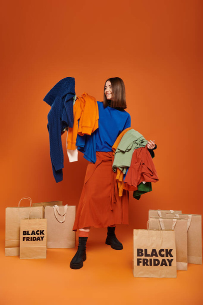 cheerful woman with brunette hair holding vibrant autumnal clothes near black friday shopping bags - Photo, Image