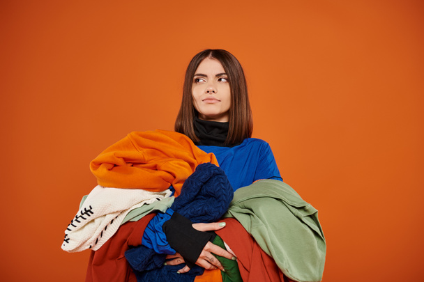 pensive young woman with brunette hair holding pile of colorful autumnal clothes, black friday - Photo, Image