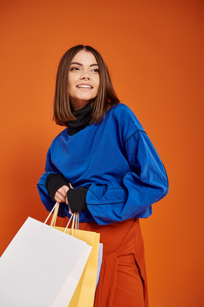 mujer alegre en ropa otoñal con estilo sosteniendo bolsas de compras el viernes negro, fondo naranja - Foto, imagen