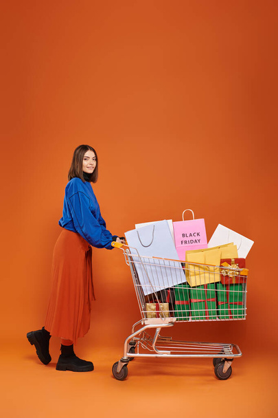 šťastná žena stojící s vozíkem plným nákupních tašek s černými páteční písmena na oranžovém pozadí - Fotografie, Obrázek