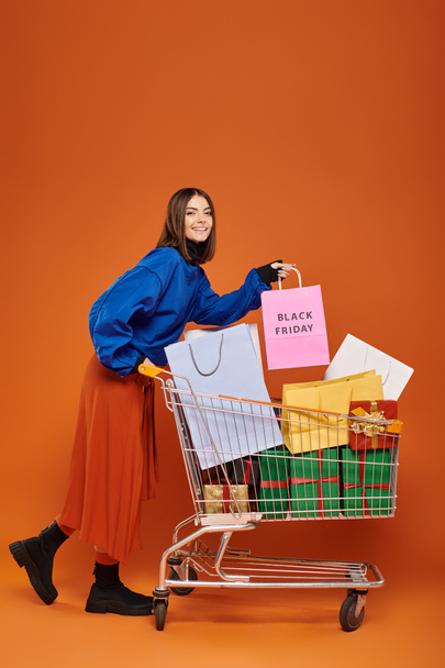 radosna kobieta stojąca z wózkiem pełnym toreb na zakupy z czarnymi, piątkowymi literami na pomarańczowym tle - Zdjęcie, obraz