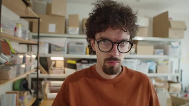 Μέτριο κοντινό πλάνο του νεαρού άνδρα με γυαλιά και casual t-shirt ποζάρουν για την κάμερα στο γραφείο της εταιρείας παροχής υπηρεσιών - Πλάνα, βίντεο