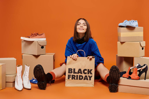 smiling brunette woman sitting with shopping bag near boxes with shoes on orange, black friday - Photo, Image