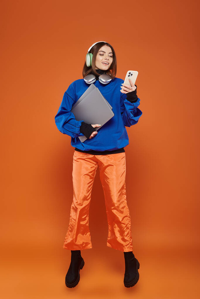 ευτυχισμένη γυναίκα με ακουστικά και φθινοπωρινή στολή χρησιμοποιώντας smartphone και κρατώντας φορητό υπολογιστή, κυβερνοχώρο Δευτέρα - Φωτογραφία, εικόνα