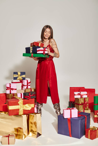 stylish brunette woman in trendy dress holding presents and looking at them, holiday gifts concept - Photo, Image