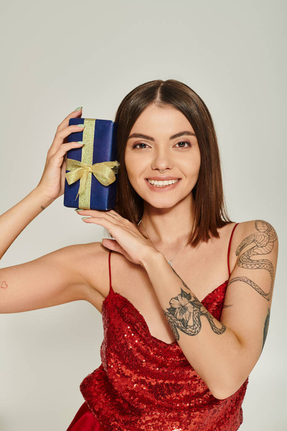dame gaie avec tatouages et piercing tenant présent près du visage souriant à la caméra, cadeaux de vacances - Photo, image