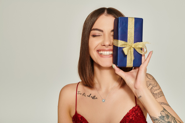 приваблива жінка тримає подарунок біля її обличчя і щиро посміхається закритими очима, святкові подарунки - Фото, зображення