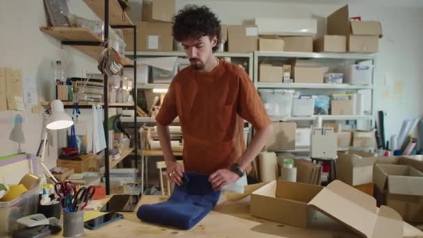 Joven trabajador masculino empacando la ropa en la caja de cartón mientras prepara el pedido para enviar al cliente en el almacén del mercado en línea - Metraje, vídeo