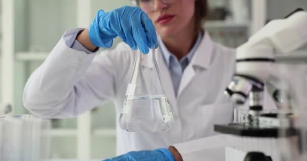 Naukowiec trzyma kolbę z przezroczystym płynem i obraca ją w dłoniach. Flask w ręce naukowca z chemicznym przezroczystym, toksycznym rozpuszczalnikiem płynnym - Materiał filmowy, wideo