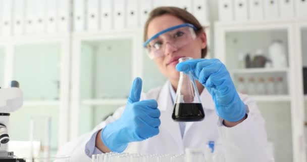 Vegyipari laboratóriumi tudós fekete folyékony olajat tart lombikban és hüvelykujjal felfelé. A kőolajtermékek minősége és jóváhagyott vizsgálata - Felvétel, videó