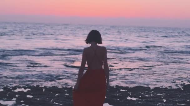 Uma jovem mulher em um vestido de verão fica em uma rocha durante o pôr do sol e olha para o mar. liberdade e beleza. - Filmagem, Vídeo