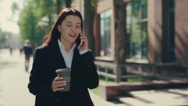 Счастливая деловая женщина в официальном костюме, идущая рядом с бизнес-центром, разговаривает по телефону с клиентами, держащими чашку эко кофе. Снаружи. Люди и технологии - Кадры, видео
