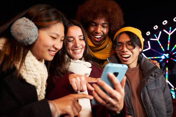 Grupa wielorasowych przyjaciół uśmiecha się patrząc i wskazując na telefon komórkowy na zewnątrz w chłodną zimową noc. Czterech studentów korzystających z technologii mobilnych stojących razem w parku rozrywki.  - Zdjęcie, obraz