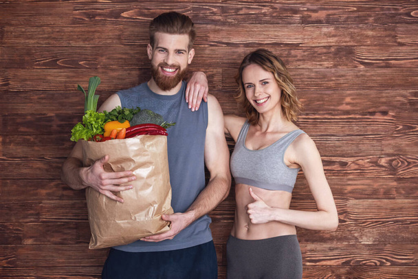 schönes junges Paar in Sportbekleidung hält eine Einkaufstasche voller gesunder Lebensmittel in der Hand, blickt in die Kamera und lächelt, auf Holzgrund - Foto, Bild