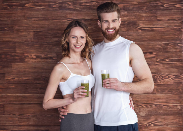 Beautiful young sports people are holding nutritious cocktails, looking at camera and smiling, on wooden background - Photo, Image
